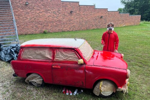 Schüler steht in Arbeitskleidung hinter dem Trabbi. Der Trabbi ist rot angesprüht.